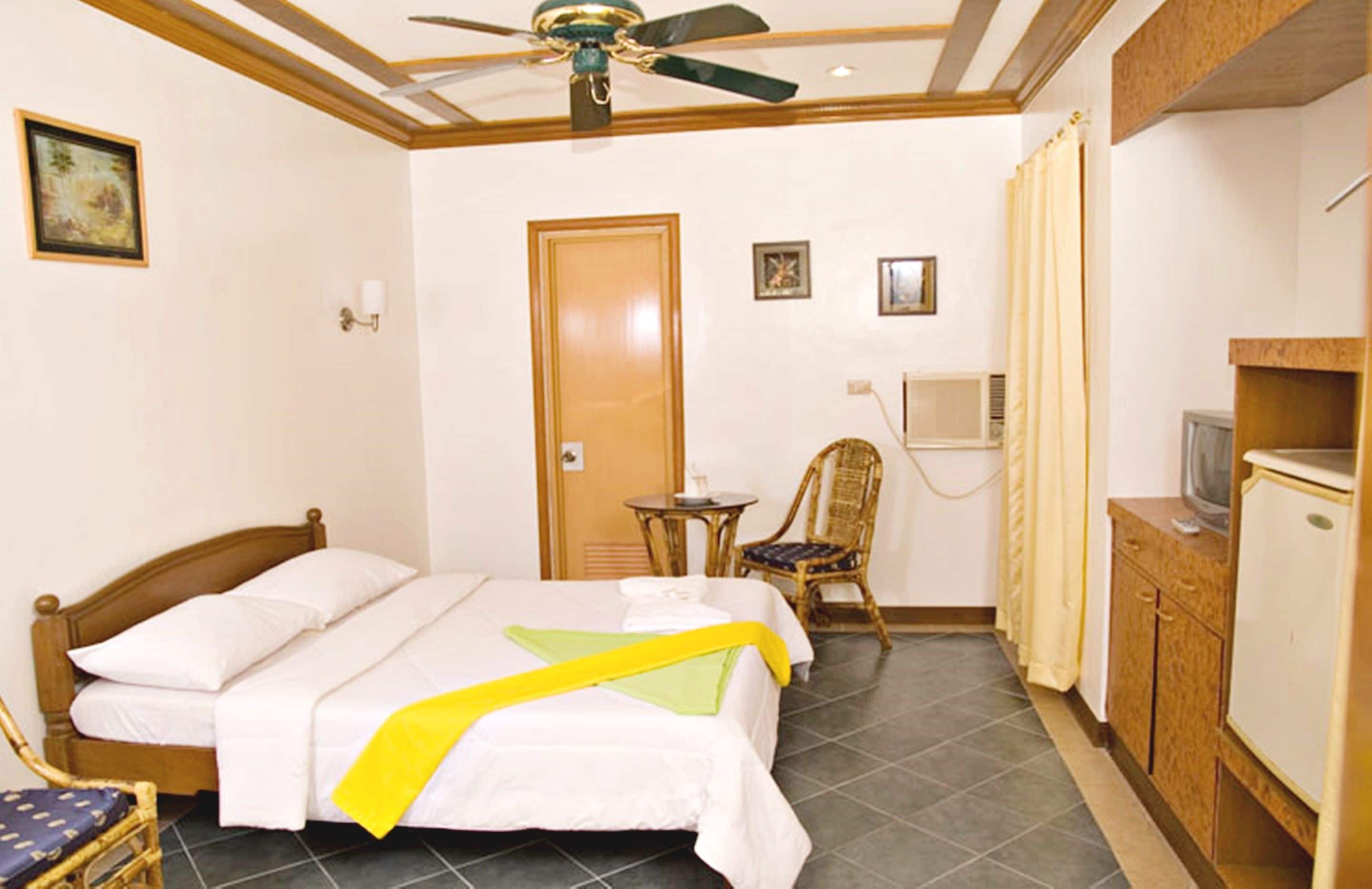 Une chambre d’hôtel équipée d’un lit, d’une chaise, d’une petite table, d’un téléviseur et d’un ventilateur de plafond.