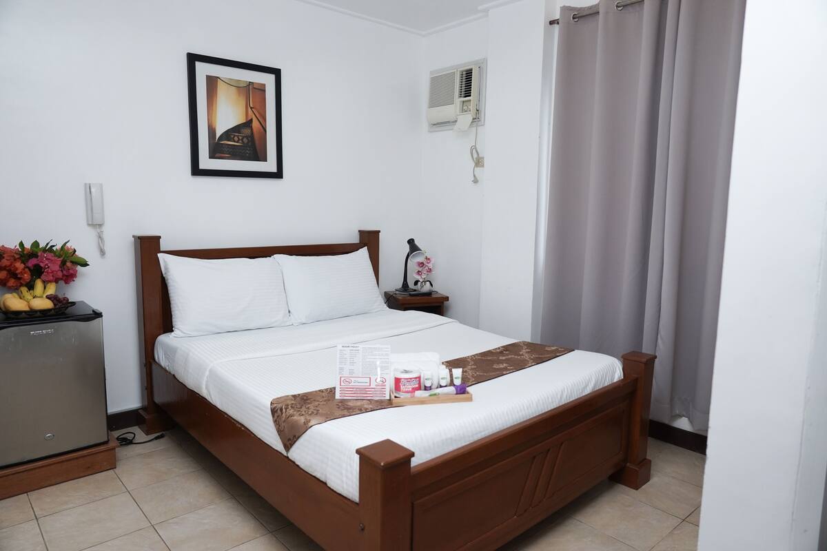 A neatly made bed with a wooden frame, a bedside table with a lamp, a refrigerator, and a wall-mounted air conditioner.