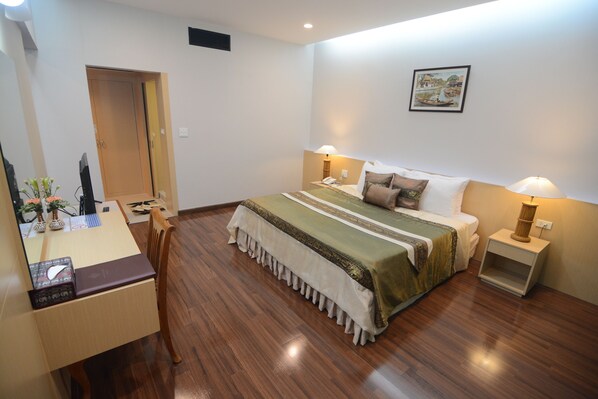 A hotel room with a wooden floor, a bed with a green bedspread, two bedside lamps, a desk with a chair, and a door. - Charoen Hotel (Udon Thani)