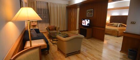 Habitación de hotel con sofá, sillón y una pequeña mesa con lámpara. Hay un televisor en un mueble bajo y una cama con almohadas.