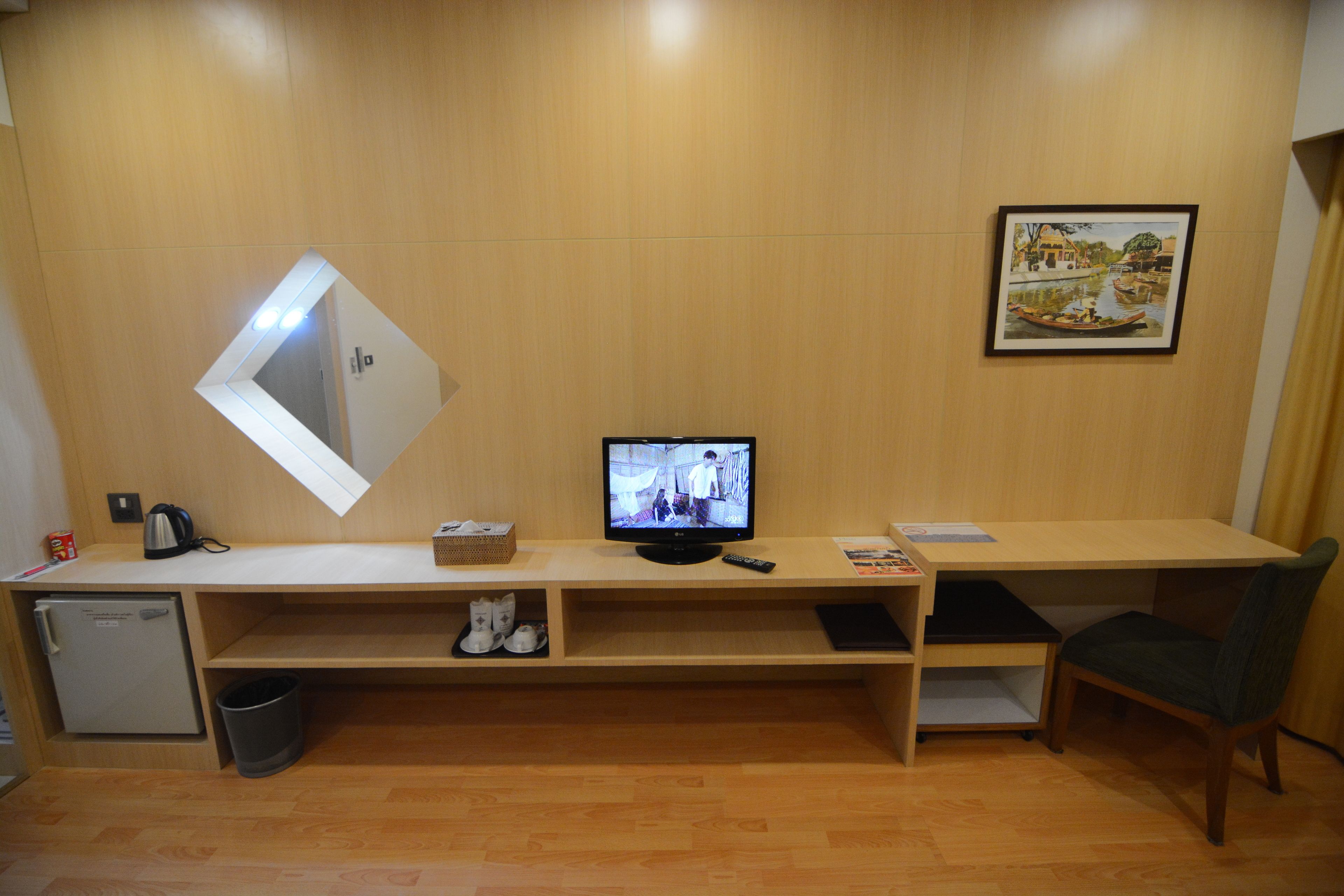 A hotel room with a wooden desk, a television, a small refrigerator, a trash bin, a chair, and a framed picture on the wall.