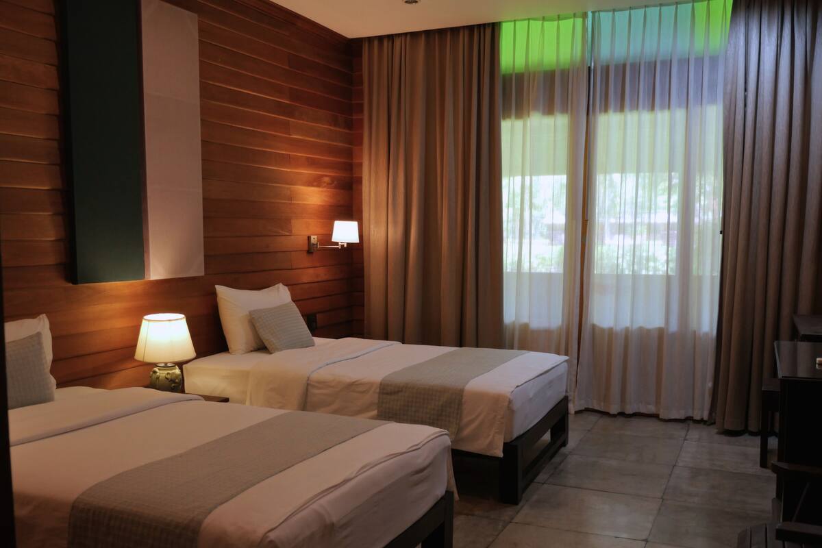 Habitación de hotel con dos camas, una mesita de noche con lámpara, cabecero de madera, ventana con cortinas traslúcidas y vista a zonas verdes.