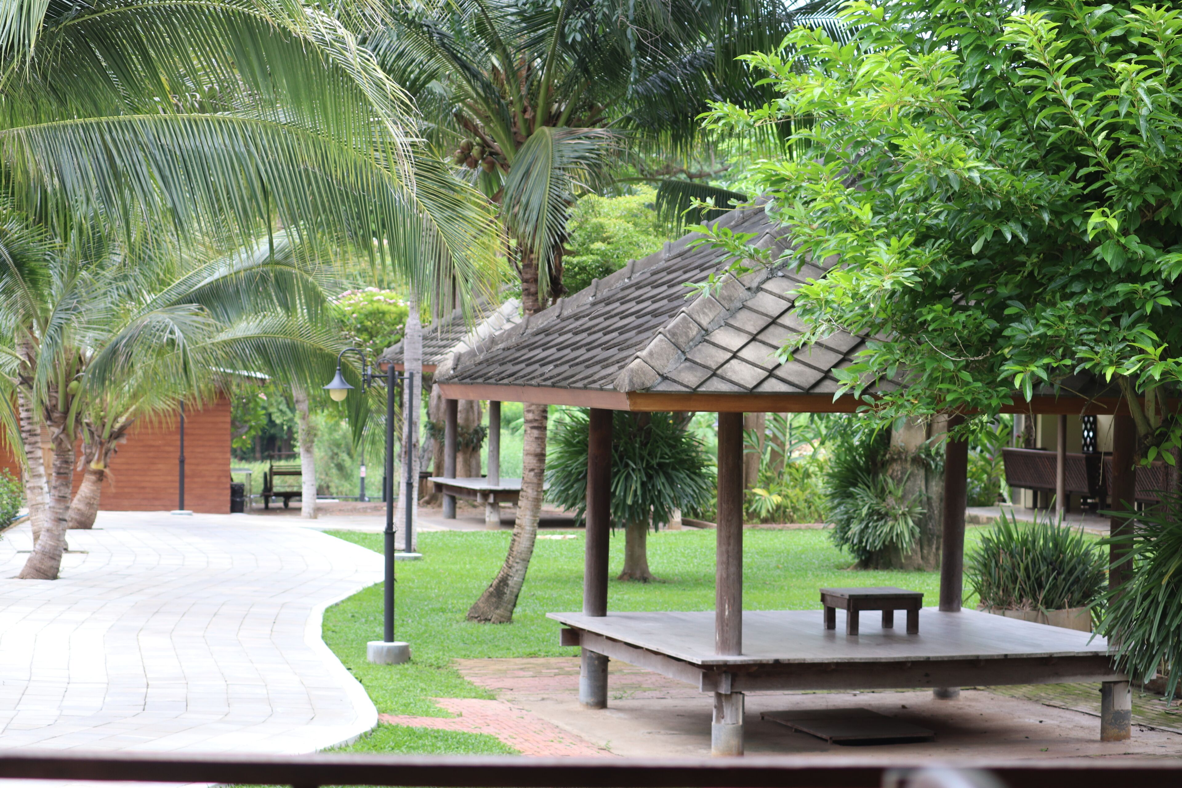 A covered outdoor seating area with benches, surrounded by lush greenery and palm trees.