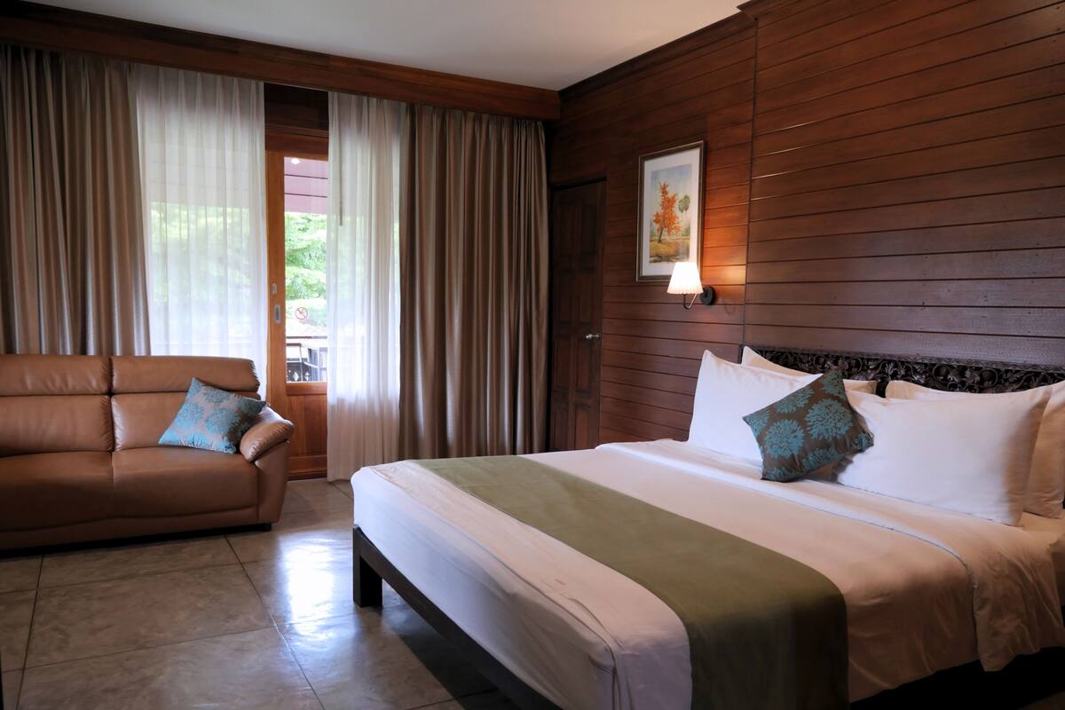 Una habitación de hotel con cama, sofá, cortinas, una lámpara fijada a la pared y un cuadro enmarco.