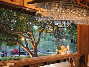 Un restaurante con terraza al aire libre, vistas a un cuerpo de agua, árboles y una estructura de madera con una lámpara colgante.