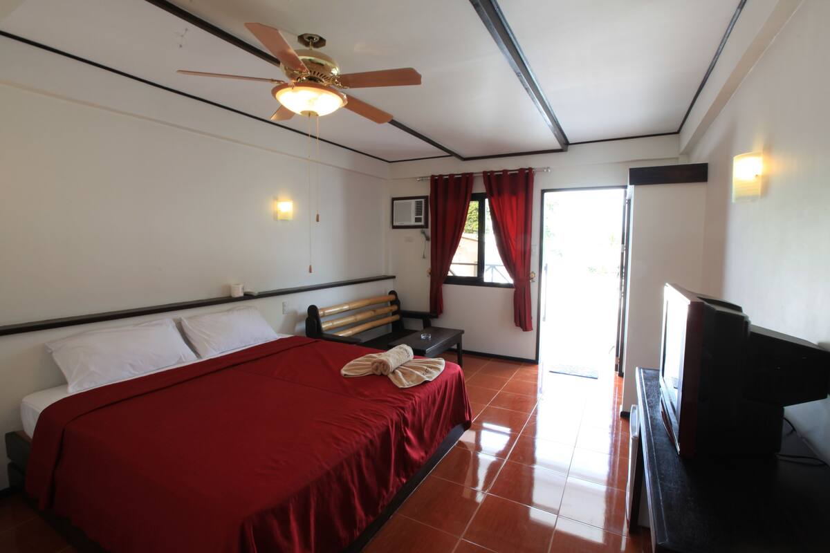 A hotel room with a bed, a ceiling fan, a television, and a window with curtains.