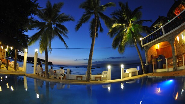 A poolside area with palm trees, a view of the sea, and a building with a balcony. - Mermaid Resort and Dive Center (Puerto Galera)