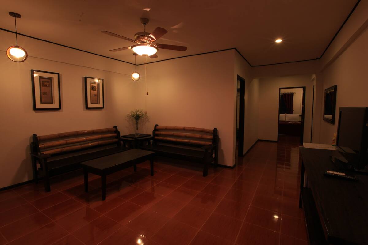 A room with a ceiling fan, two framed pictures on the wall, a wooden bench, a small table, and a television on a stand.