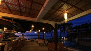 An outdoor dining area with tables and chairs, a bar with bottles and glasses, and a view of the ocean at dusk. - Mermaid Resort and Dive Center (Puerto Galera)