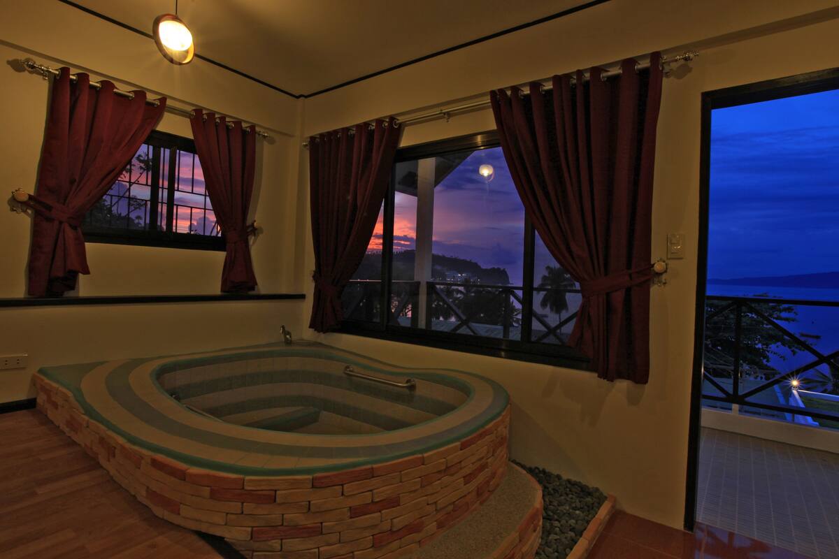 A spa room with a stone hot tub, large windows with curtains, and a view of the sunset.