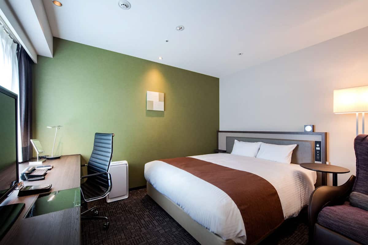 A hotel room with a bed, a desk with a chair, a lamp, and a small table.