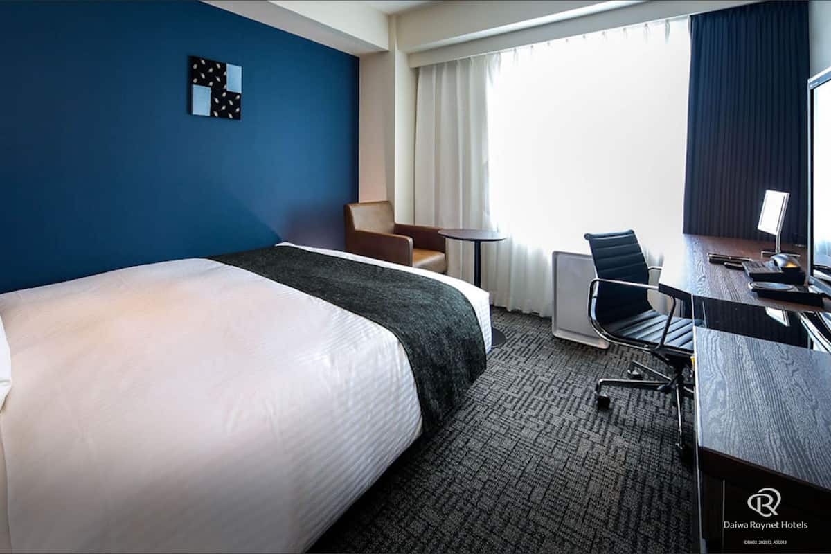 A hotel room with a large bed, a desk with a chair, a small table, and a window with curtains.