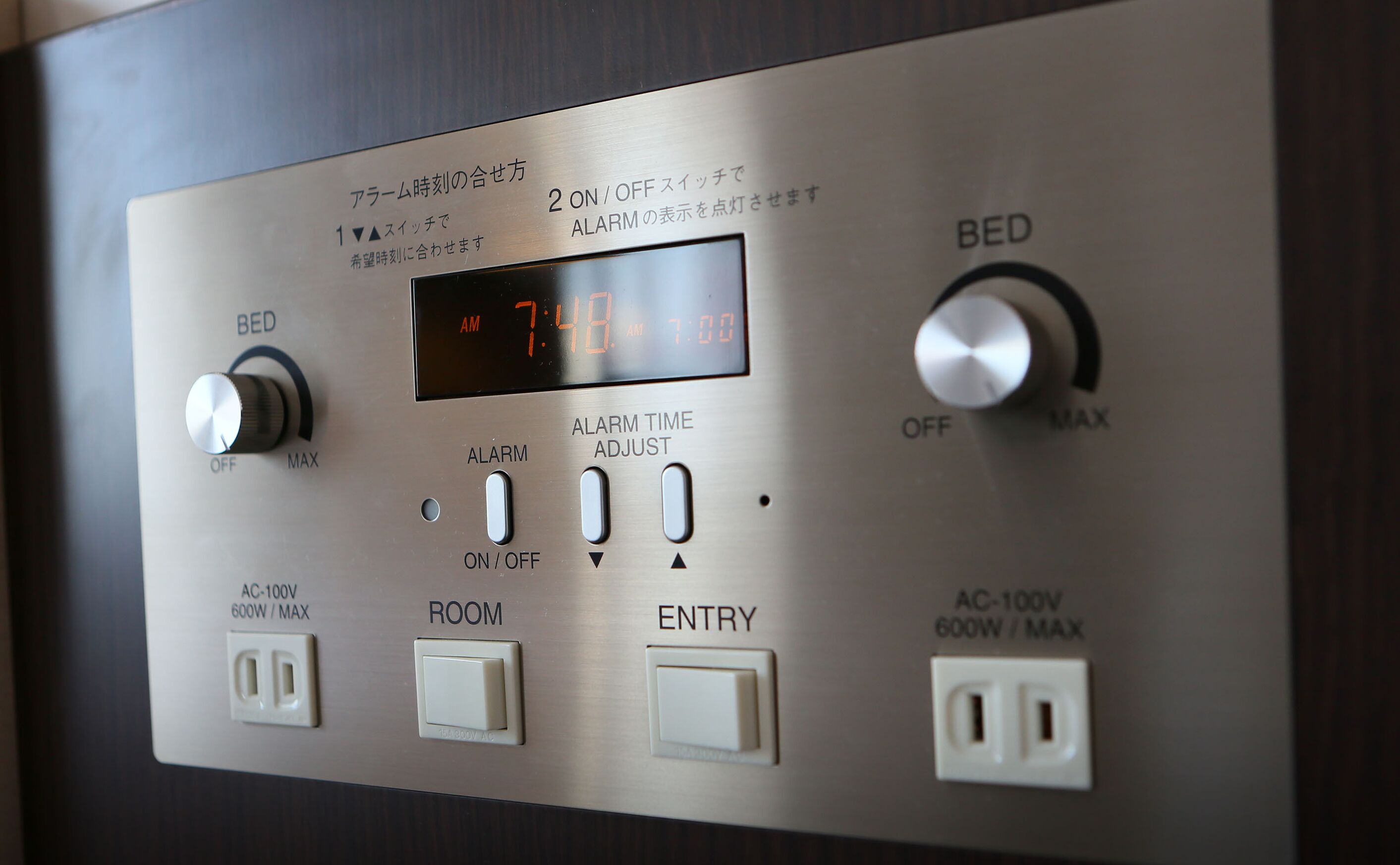 Een bedieningspaneel met een digitale klok waarop 7:48 aangegeven staat, alarminstellingen en stopcontacten met de aanduiding AC-100V 600W MAX.