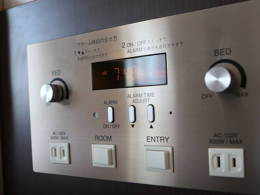 Een bedieningspaneel met een digitale klok waarop 7:48 aangegeven staat, alarminstellingen en stopcontacten met de aanduiding AC-100V 600W MAX.