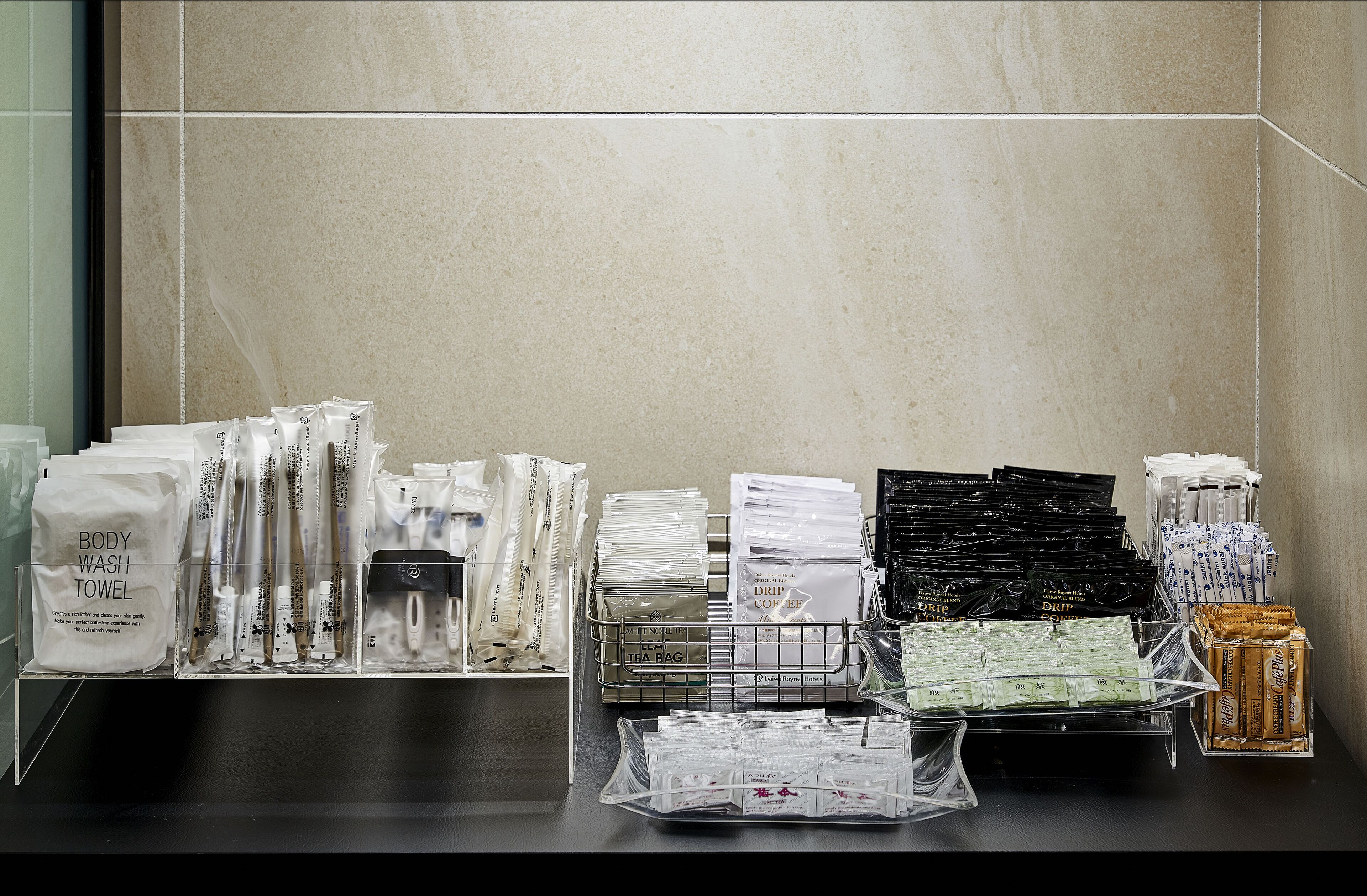 Un baño de hotel con artículos de aseo organizados en estantes y encimeras, incluyendo gel de ducha, toallas y café.