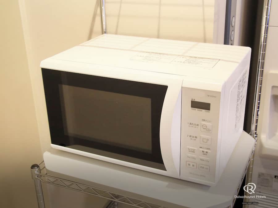 Un four à micro-ondes blanc, doté d’un écran numérique et d’un panneau de commande, est posé sur une étagère en métal dans une chambre d’hôtel.