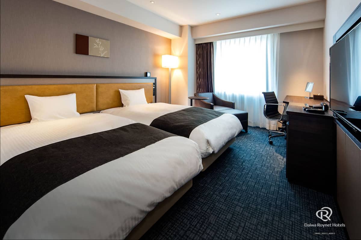 A hotel room with two beds, a desk, a chair, a TV, and a window with curtains.