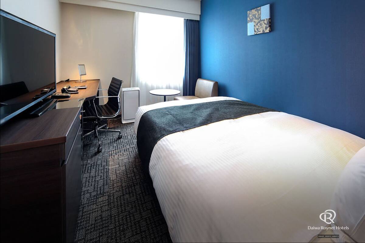 A hotel room with a bed, a desk with a computer, a chair, and a television.