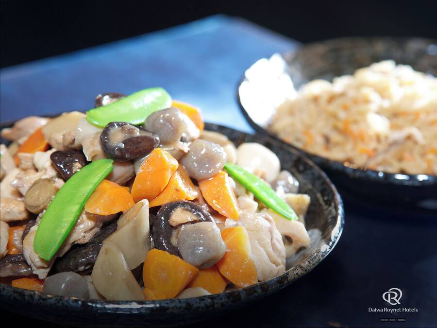 A close-up of a dish with various vegetables and meat, and a bowl of rice in the background.