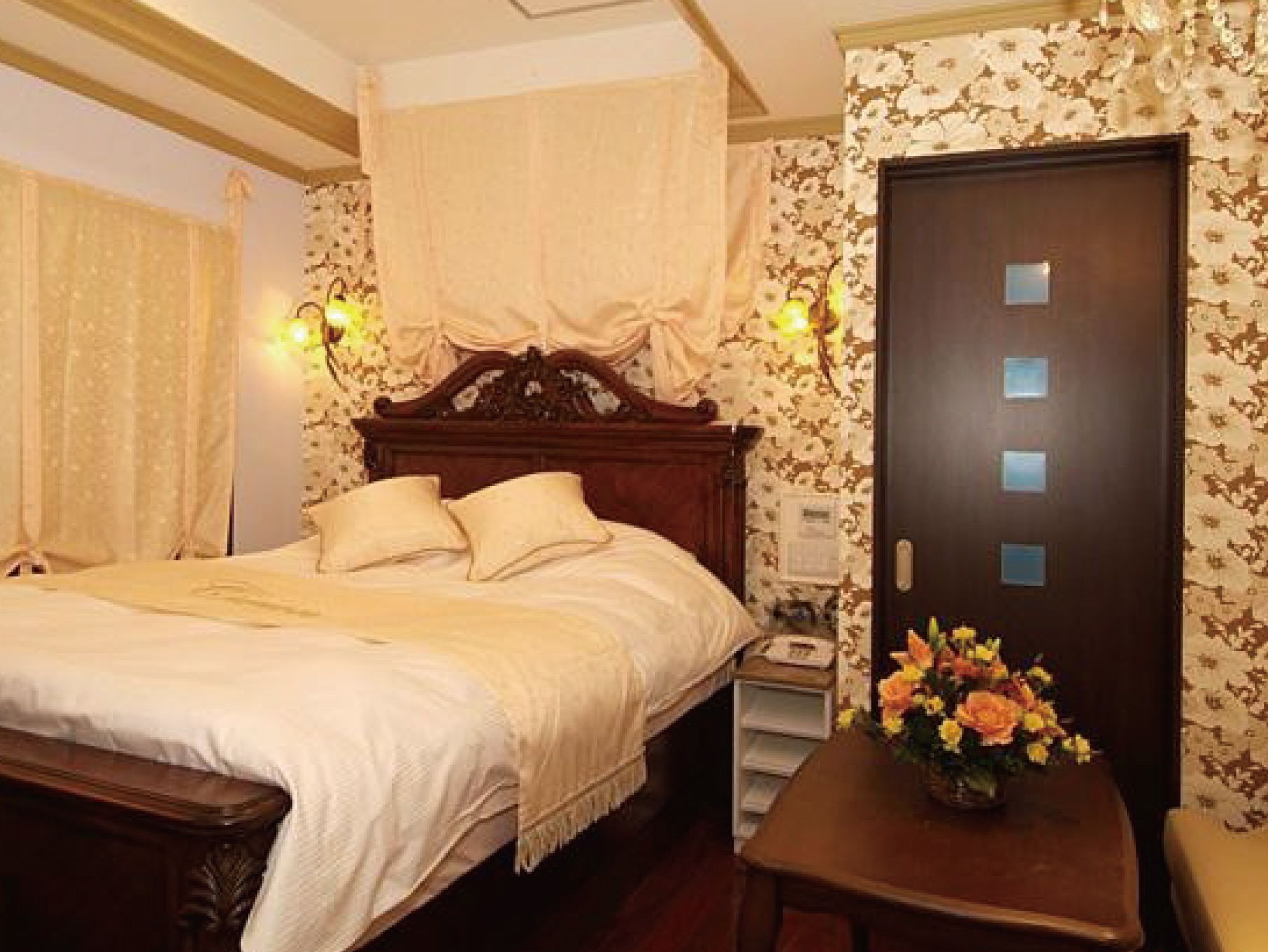A neatly made bed with a wooden headboard, a bedside table with a vase of flowers, and a door with a light switch panel.