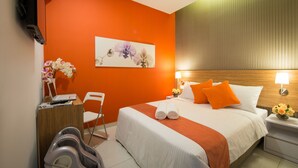 Een hotelkamer met een oranje accentmuur, een bed met wit en oranje beddengoed, een bureau met een laptop, een stoel en een massagestoel.