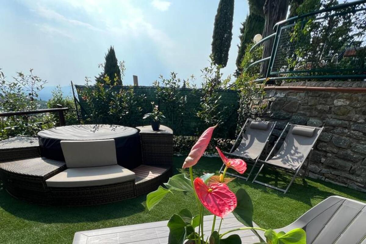 Een terras met een ronde tafel en stoelen, een kleine potplant met roze bloemen, en een stenen muur op de achtergrond.