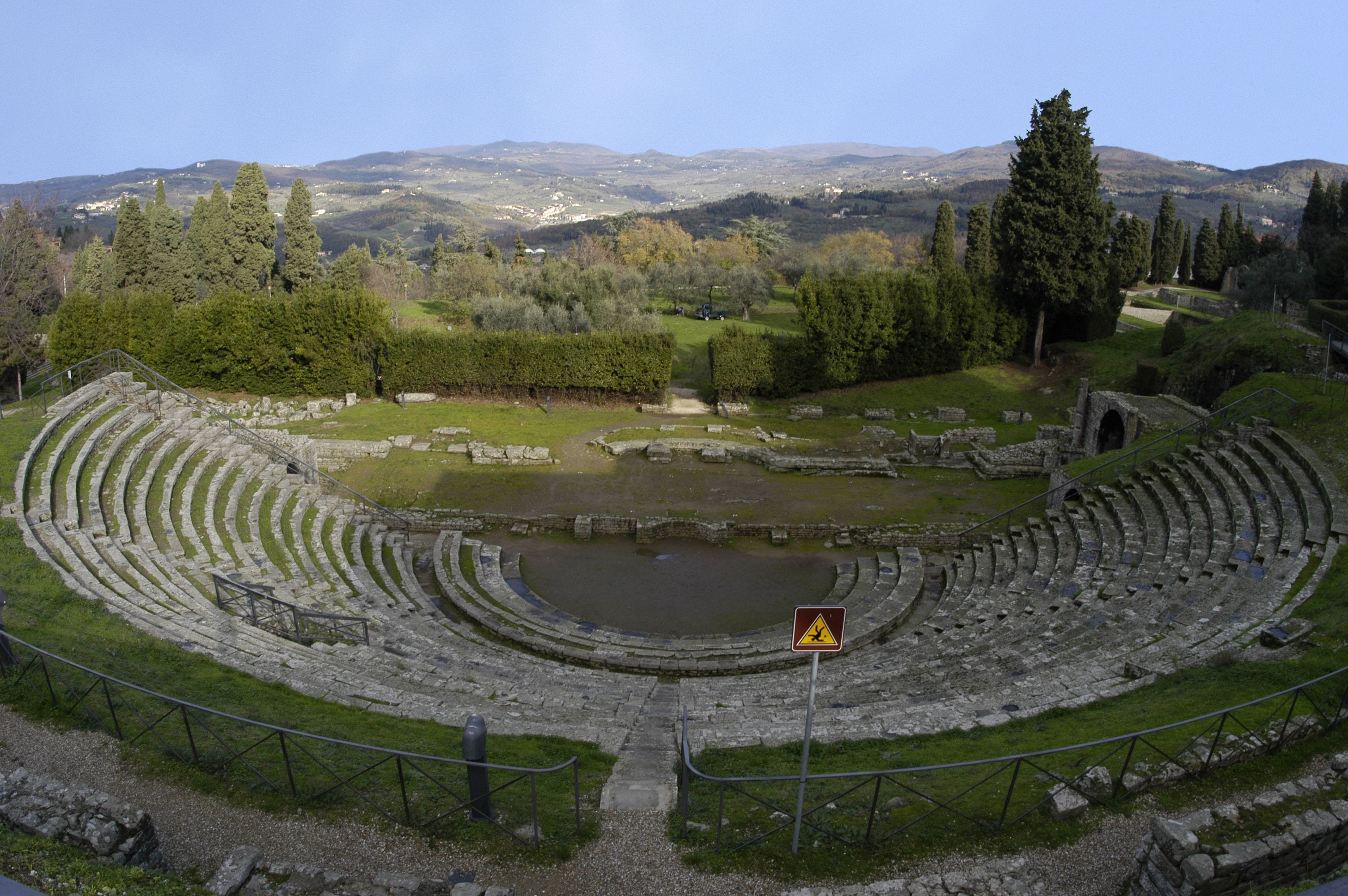 An ancient amphitheater with stone seating and a central stage, surrounded by trees and hills.