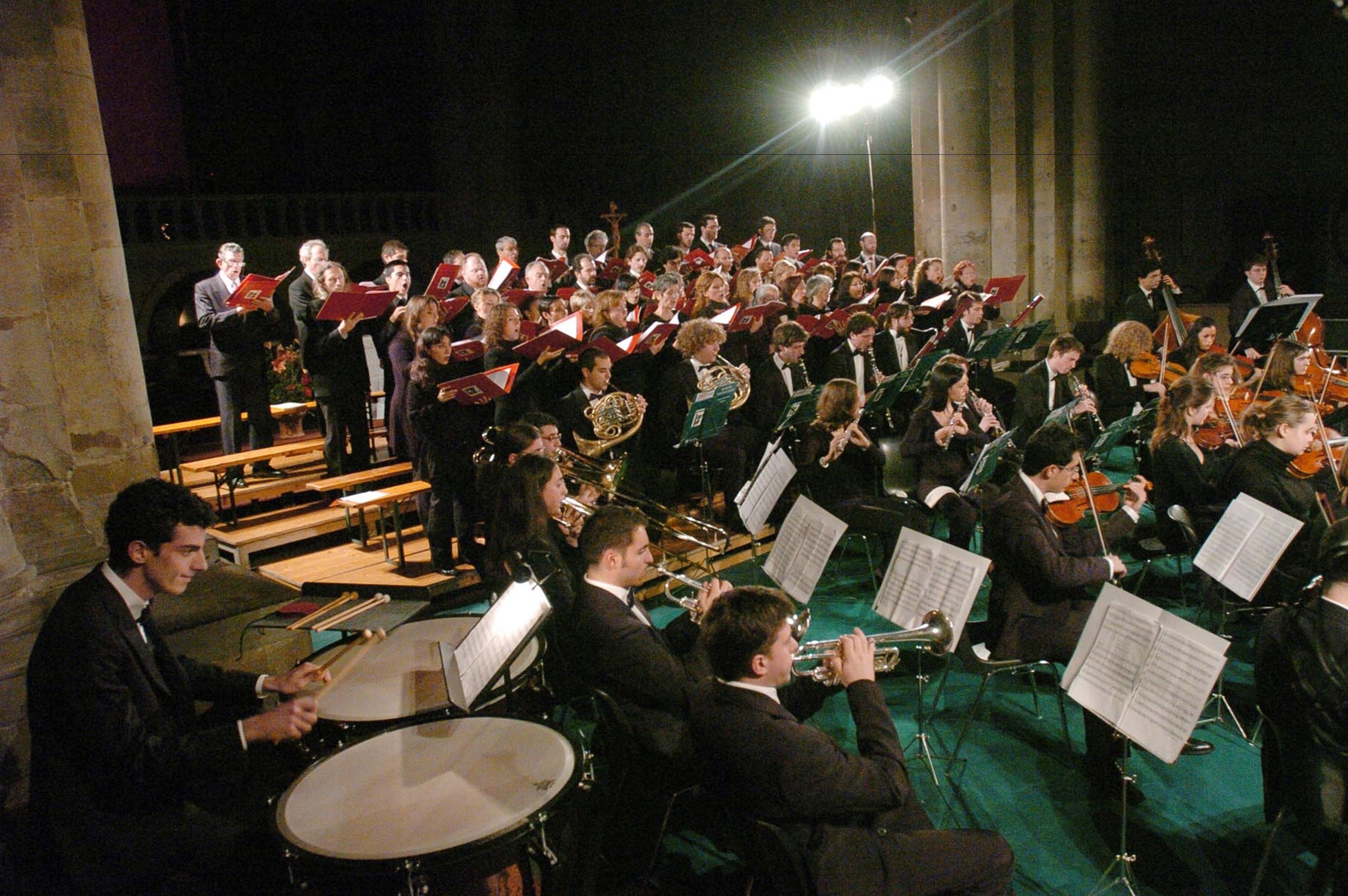 A large orchestra performing on stage with musicians seated and standing, and music stands visible.
