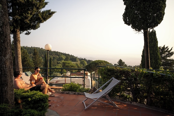 A terrace with a lounge chair, a person sitting on a bench, and a view of a hilly landscape.