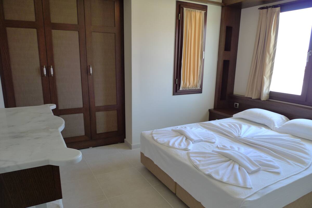Ein Schlafzimmer mit einem Bett, einem Holzschrank, einem Badezimmer mit weißer Ablage und einem Fenster mit Vorhängen.