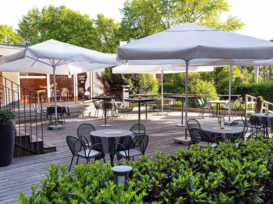 An outdoor patio area with tables and chairs, covered by umbrellas, and surrounded by greenery.