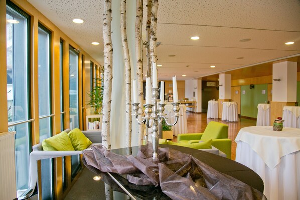 Un hall d’hôtel moderne avec une table en verre, un chandelier, un fauteuil vert et une table recouverte d’une nappe blanche.