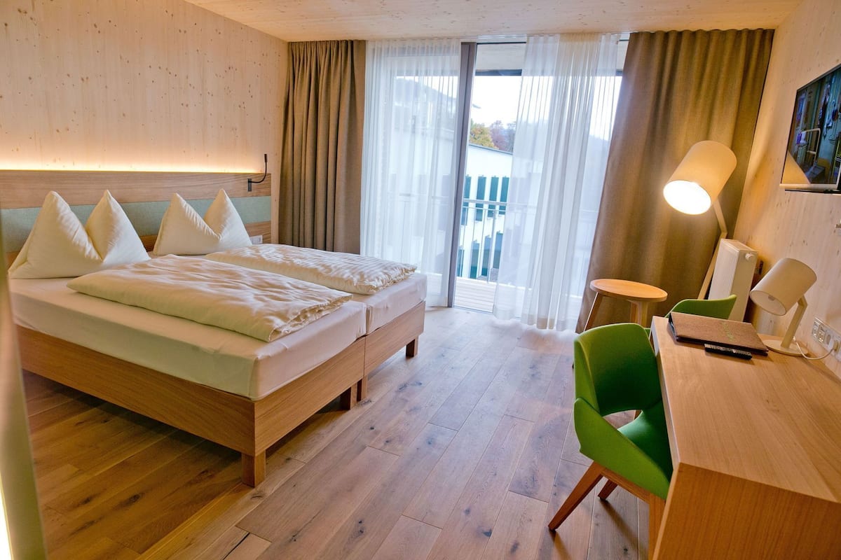 Een hotelkamer met een houten bed, een bureau met lamp en uitzicht op het omliggende landschap door een groot raam.