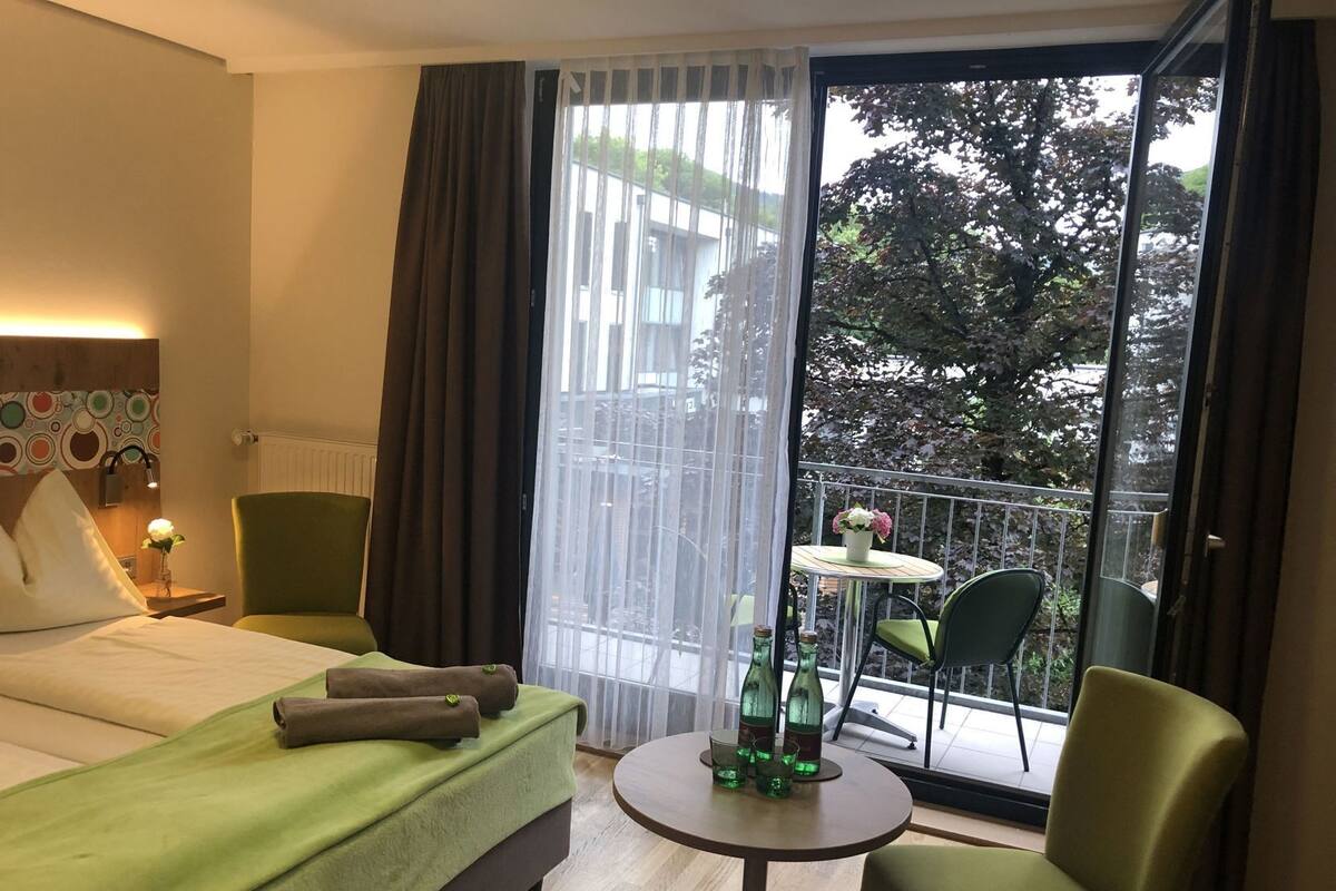 Een hotelkamer met een bed, een stoel, een tafeltje en een balkon met uitzicht op bomen en gebouwen.