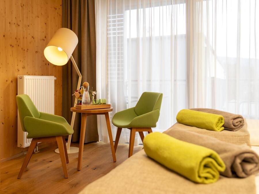 Ett hotellrum med träväggar, en säng med hoprullade handdukar, två gröna stolar, ett litet bord med en lampa och ett fönster med tunna, genomskinliga gardiner.