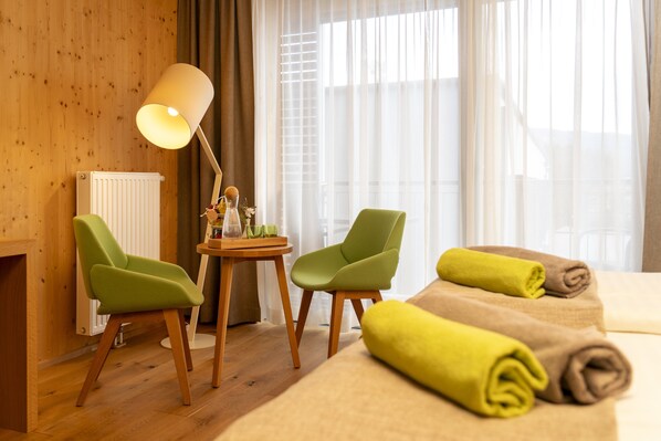 Une chambre d’hôtel aux murs en bois, un lit avec des serviettes pliées, deux fauteuils verts, une petite table avec une lampe et une fenêtre ornée de rideaux légers.
