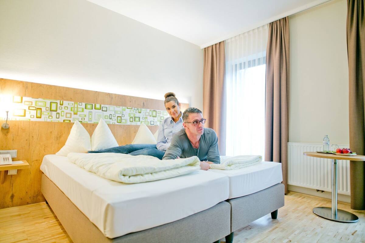 Een tweepersoonsbed met hoofdeinde, een nachtkastje met lamp, een klein rond tafeltje met fles en fruit, en een raam met gordijnen.