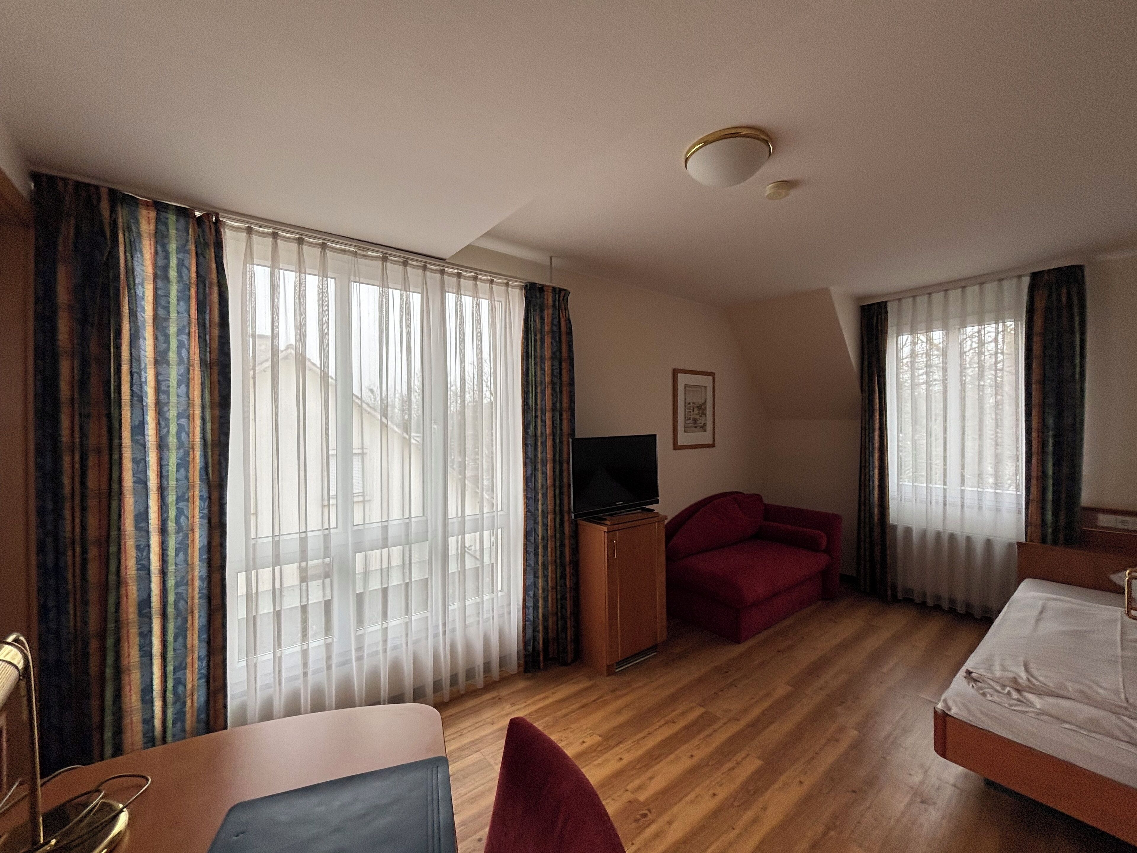 Una habitación de hotel con piso de madera, una cama, un televisor, un sofá rojo y cortinas.