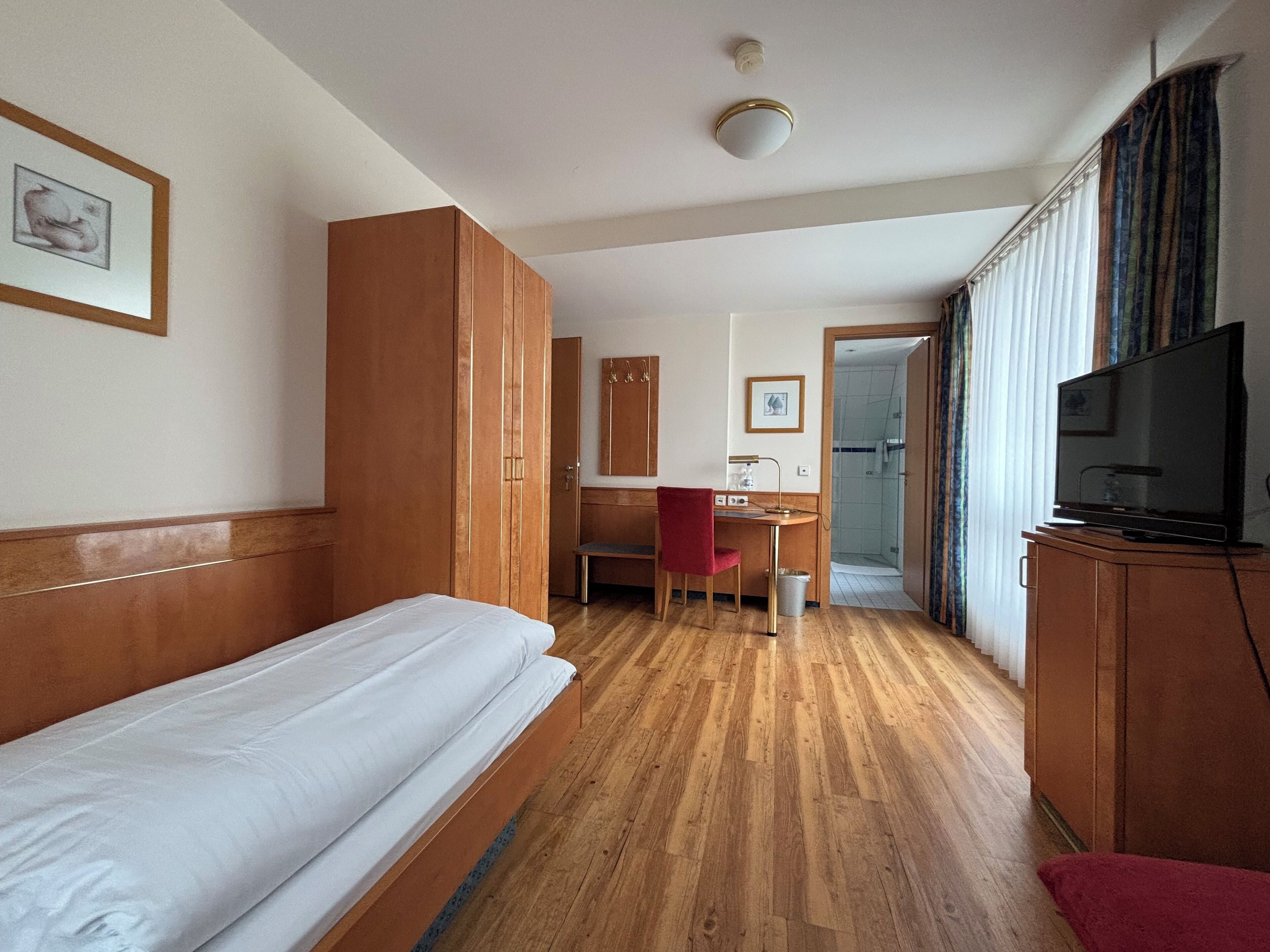 A hotel room with a single bed, a desk with a chair, a television, and a wardrobe.