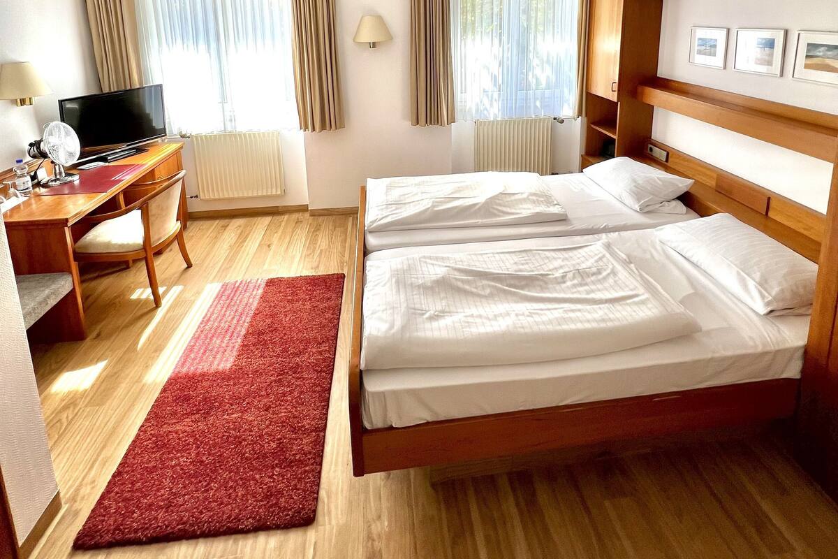 Ein Hotelzimmer mit einem hölzernen Bett, einem Schreibtisch mit Computer, einem Fernseher und einem roten Teppich.