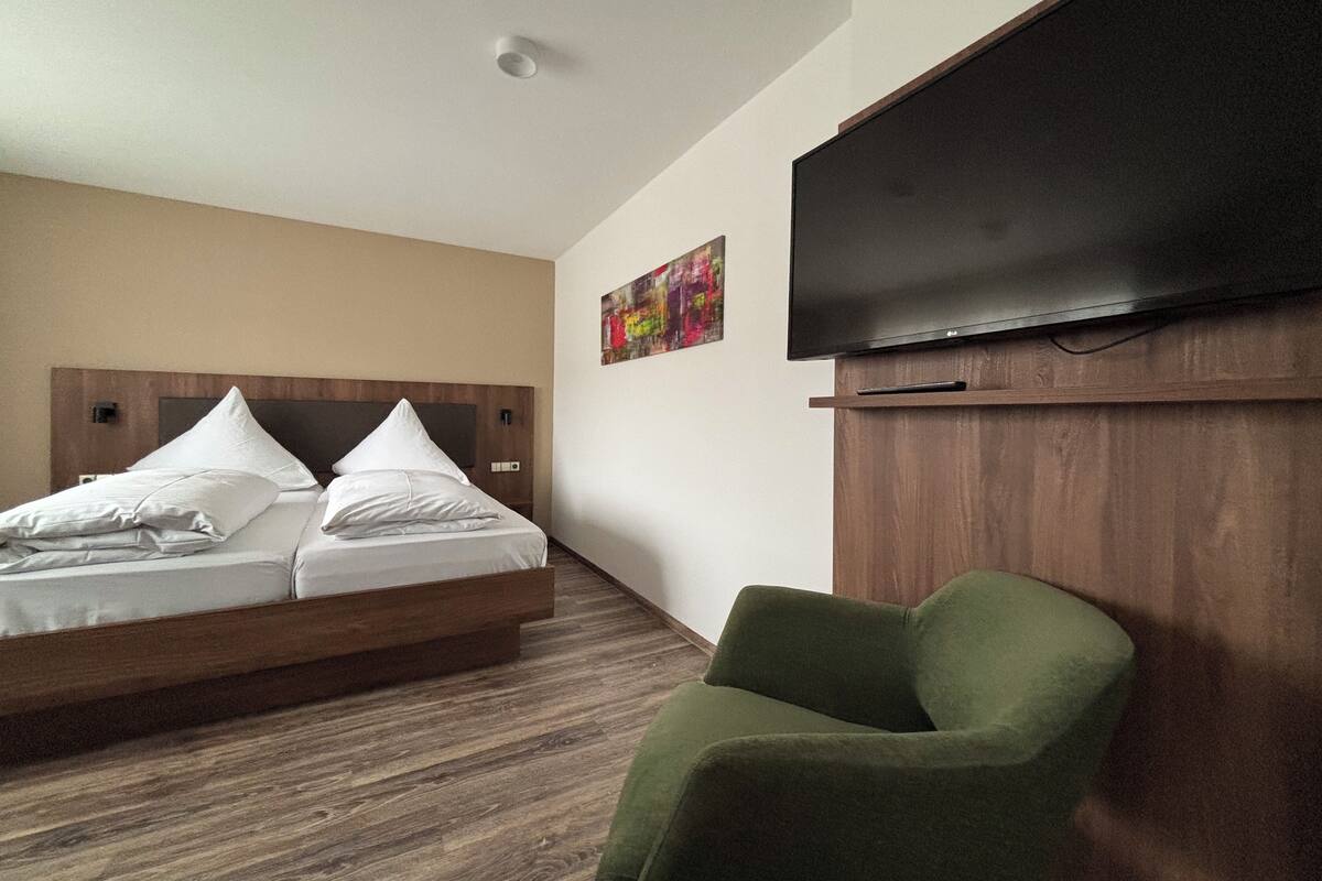 Ein Hotelzimmer mit einem Bett, einem grünen Sessel, einem Flachbildfernseher und einem hölzernen Kopfteil.