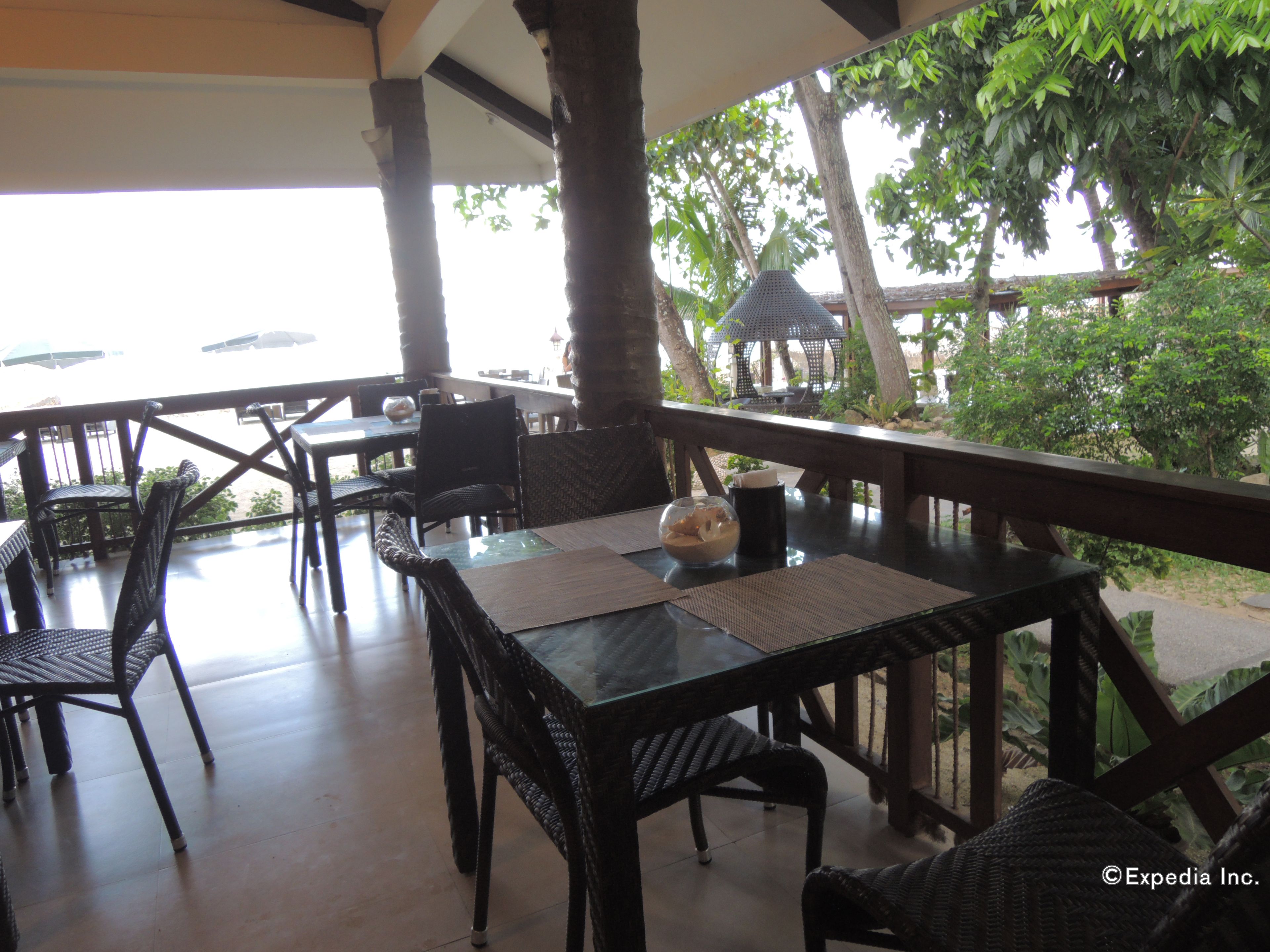 An outdoor dining area with wicker chairs and tables, surrounded by trees and a view of the sea.