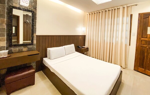 A hotel room with a bed, a wooden headboard, a nightstand, a mirror, a wooden door, and a window with curtains.