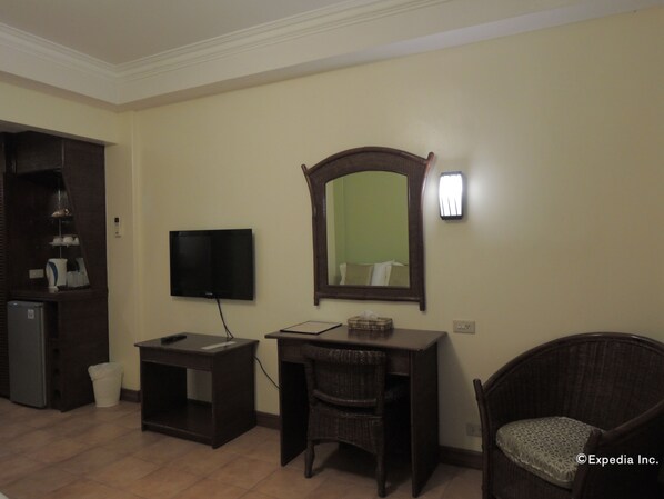 A room with a television, a desk, a wicker chair, and a mirror on the wall.