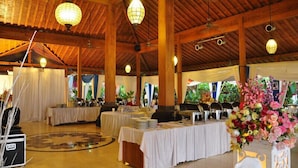 Une vaste salle dotée de hauts plafonds, de poutres en bois et de lustres. Les tables sont dressées avec des nappes blanches et décorées de compositions florales.