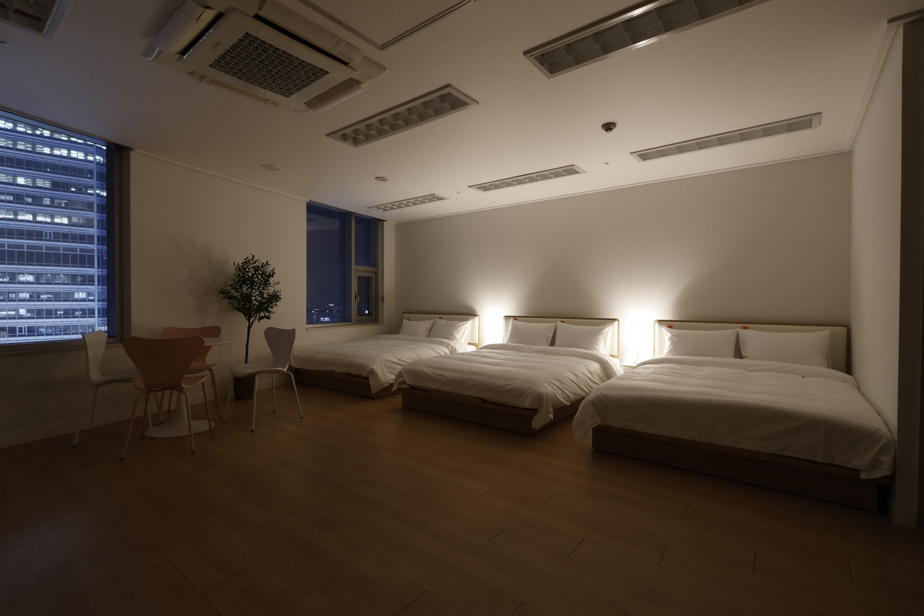 A hotel room with two beds, a desk, chairs, and a large window with a city view.