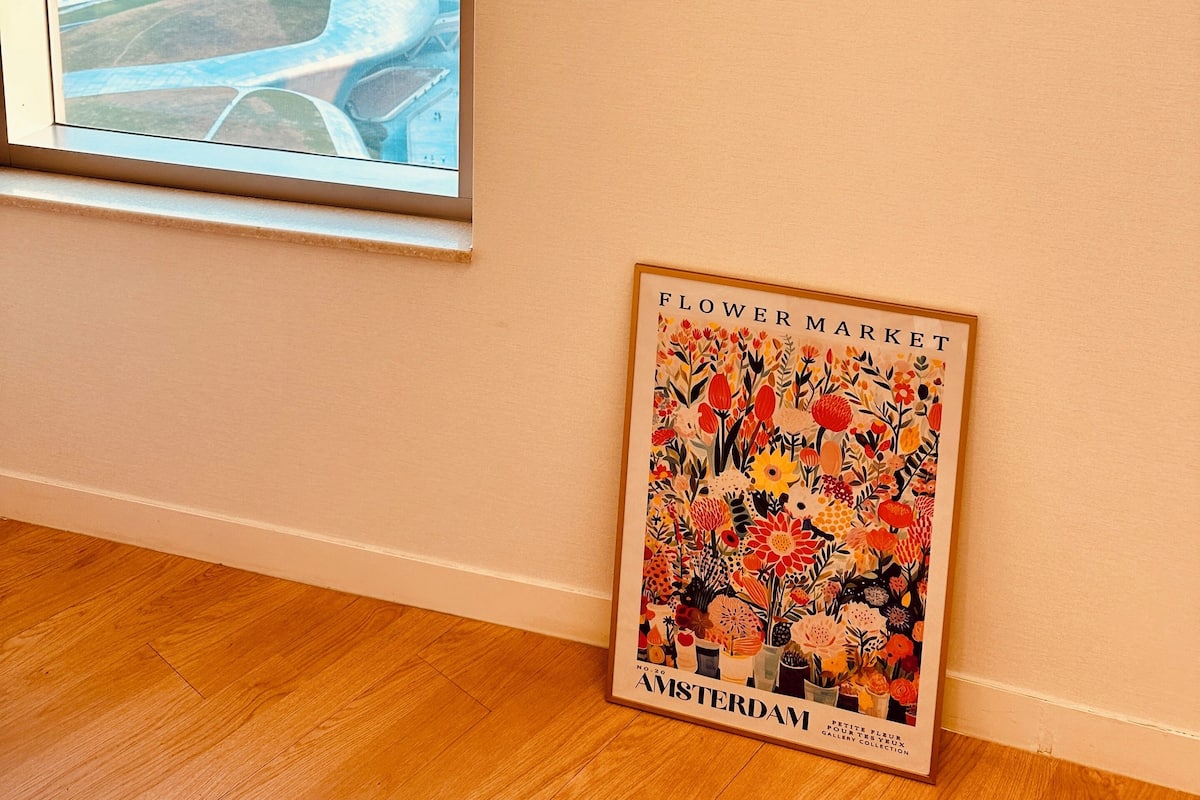 Une pièce avec une fenêtre donnant sur une vue de la ville, un plancher en bois et une affiche représentant le marché aux fleurs d’Amsterdam.