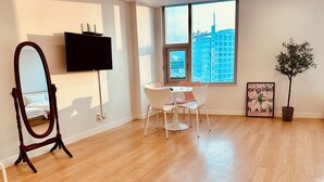 Una habitación moderna con piso de madera, un ventanal con vista a la ciudad, televisor de pantalla plana, mesa de comedor con sillas, espejo y un cuadro enmarcado.