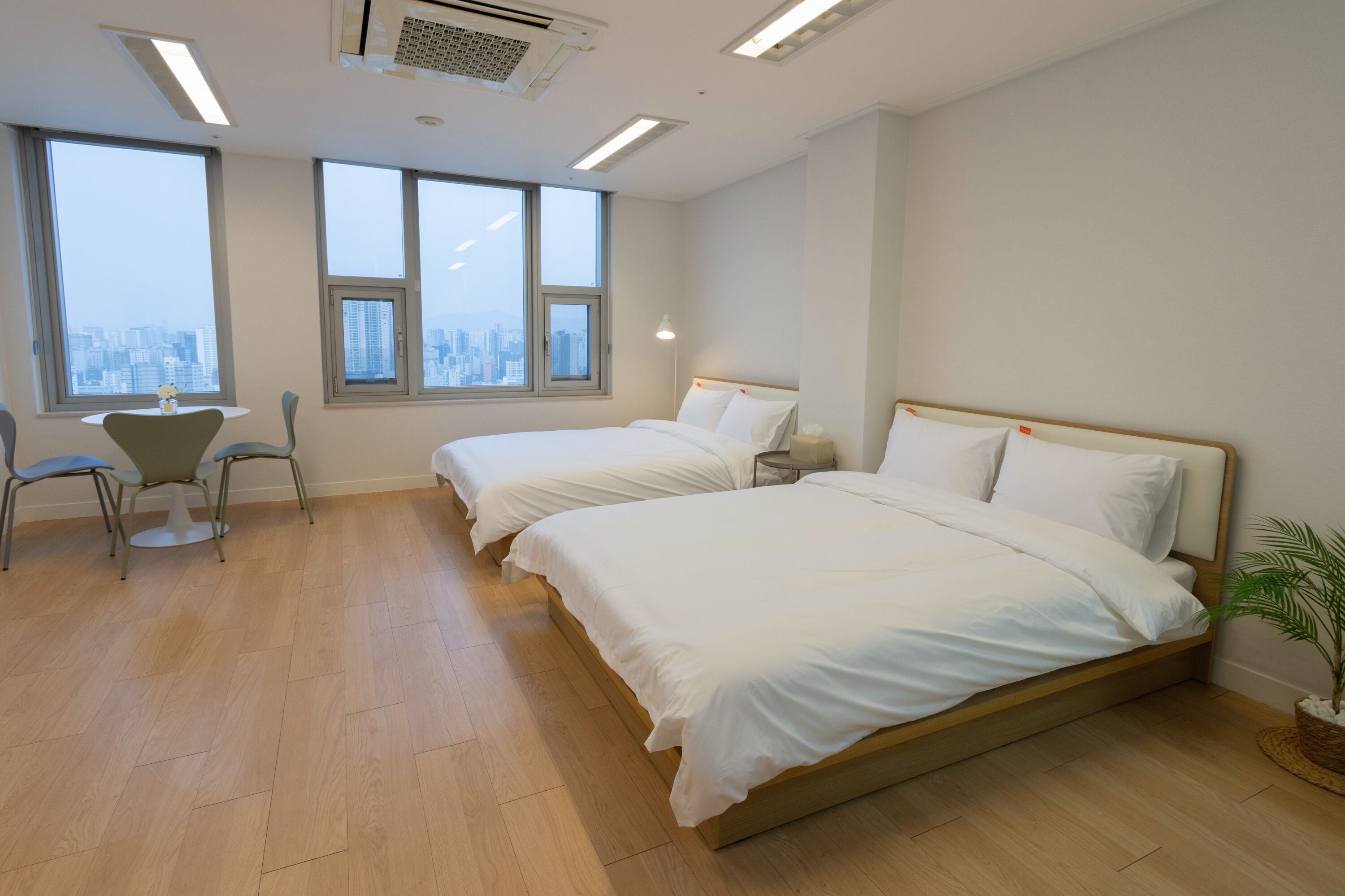 Habitación de hotel con dos camas, un escritorio y ventanas amplias con vistas a la ciudad.