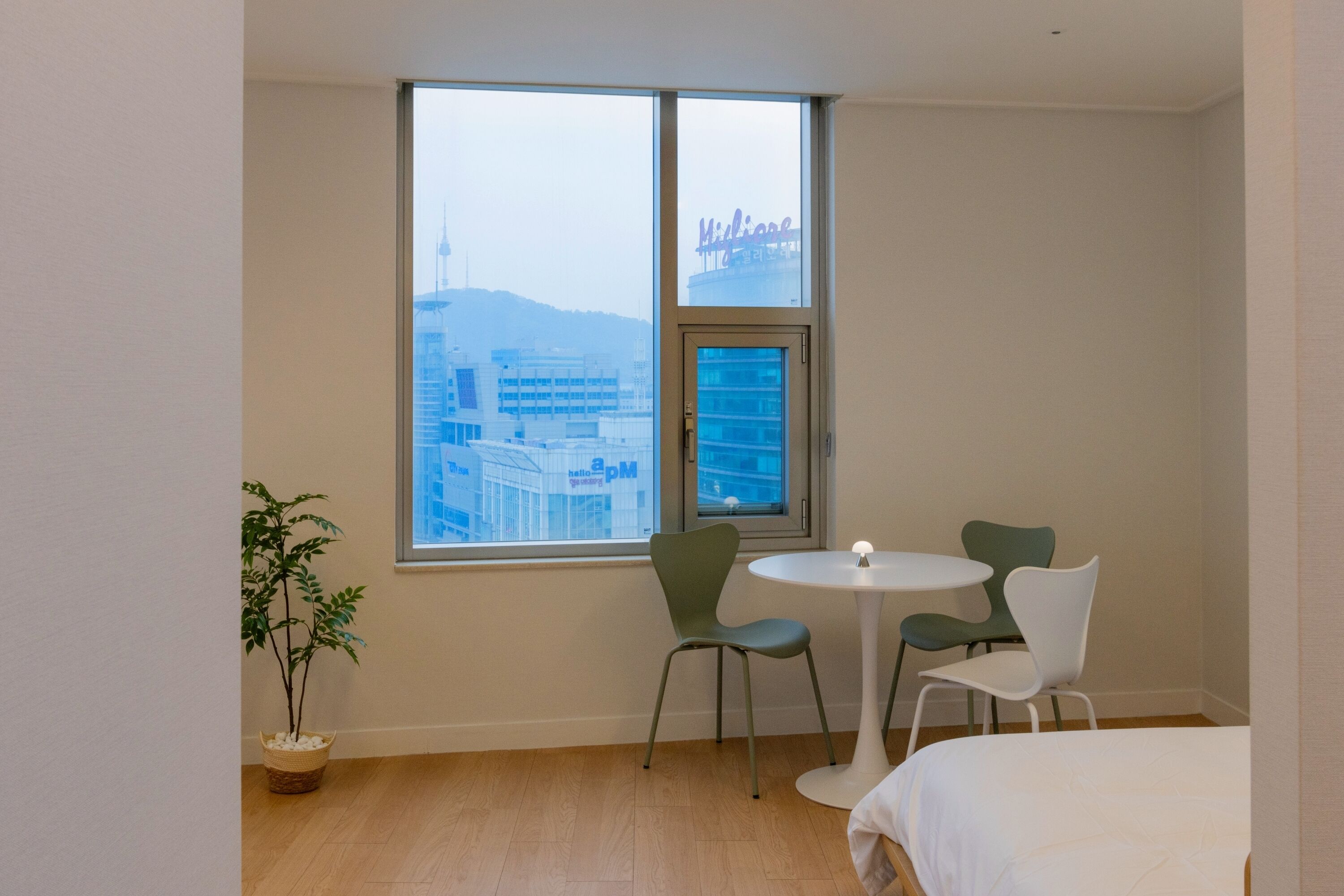 Habitación de hotel con una ventana grande, una cama, una mesita redonda pequeña, dos sillas y una planta en maceta.